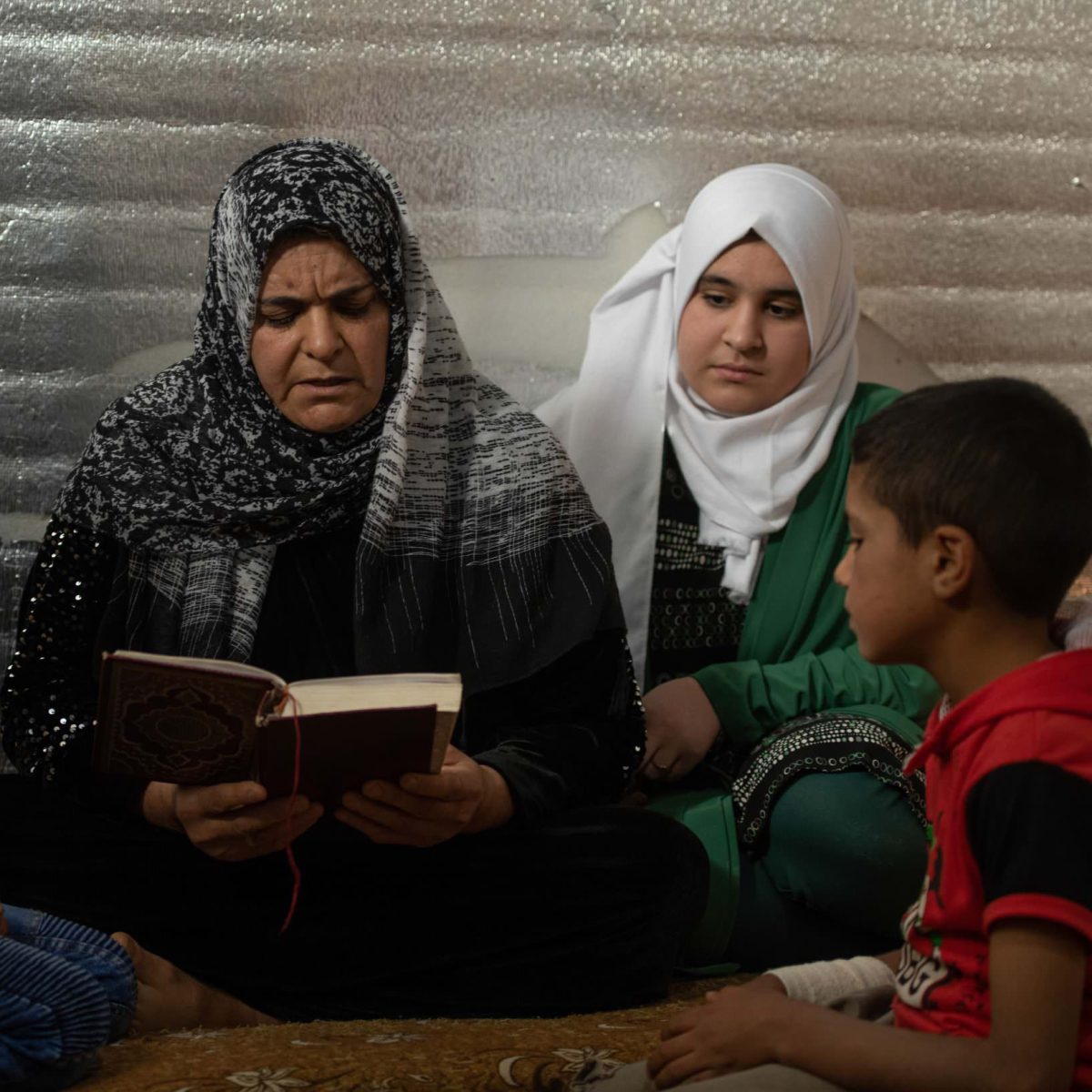Um Hadi reads the Qur’an with her three children Hadi (8), Osama (7) and Shatha (13) at home in Azraq Camp. 

During Ramadan the family spend more time praying and reading the Qur’an. Shatha says “This month of Ramadan we should treat each other in nice way and be kind too since it’s a month of forgiveness and compassion.” ; Um Hadi (47) fled her home in Homs, Syria in 2016 after her husband based away. Now living in Azraq Camp, the is the primary guardian for her three children Hadi (8), Osama (7) and Shatha (13).
 
This Ramadan is their sixth in Jordan and Um Hadi says that it its at this time of year when she misses her family back in Syria the most “I call my mom every day to check on her.”